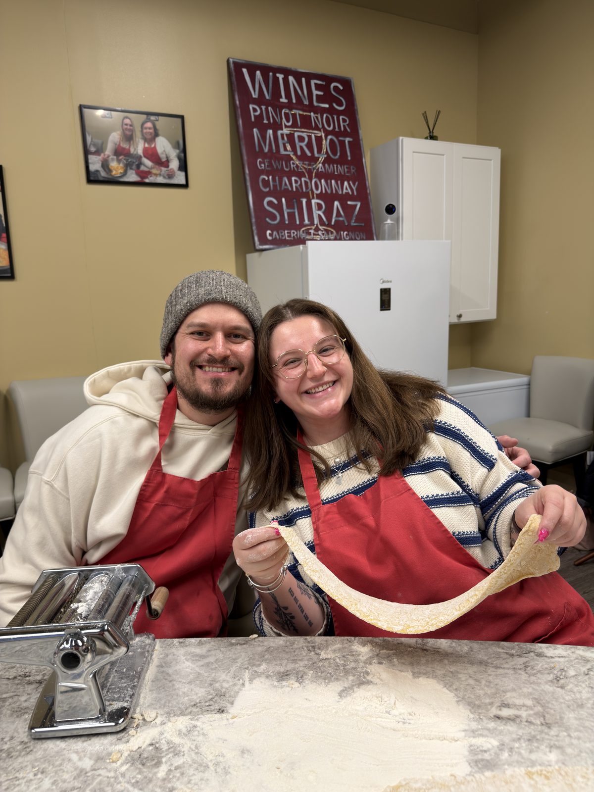 Cody and his partner in red aprons at a pasta class, holding up a sheet of fresh dough